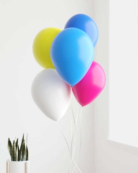 A colorful bunch of balloons in yellow, blue, pink, white, and green floats against a bright wall, with greenery nearby.