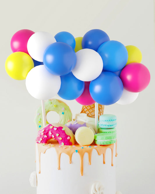 Colorful balloons in blue, pink, white, and yellow atop a cake adorned with donuts, ice cream, and macarons.