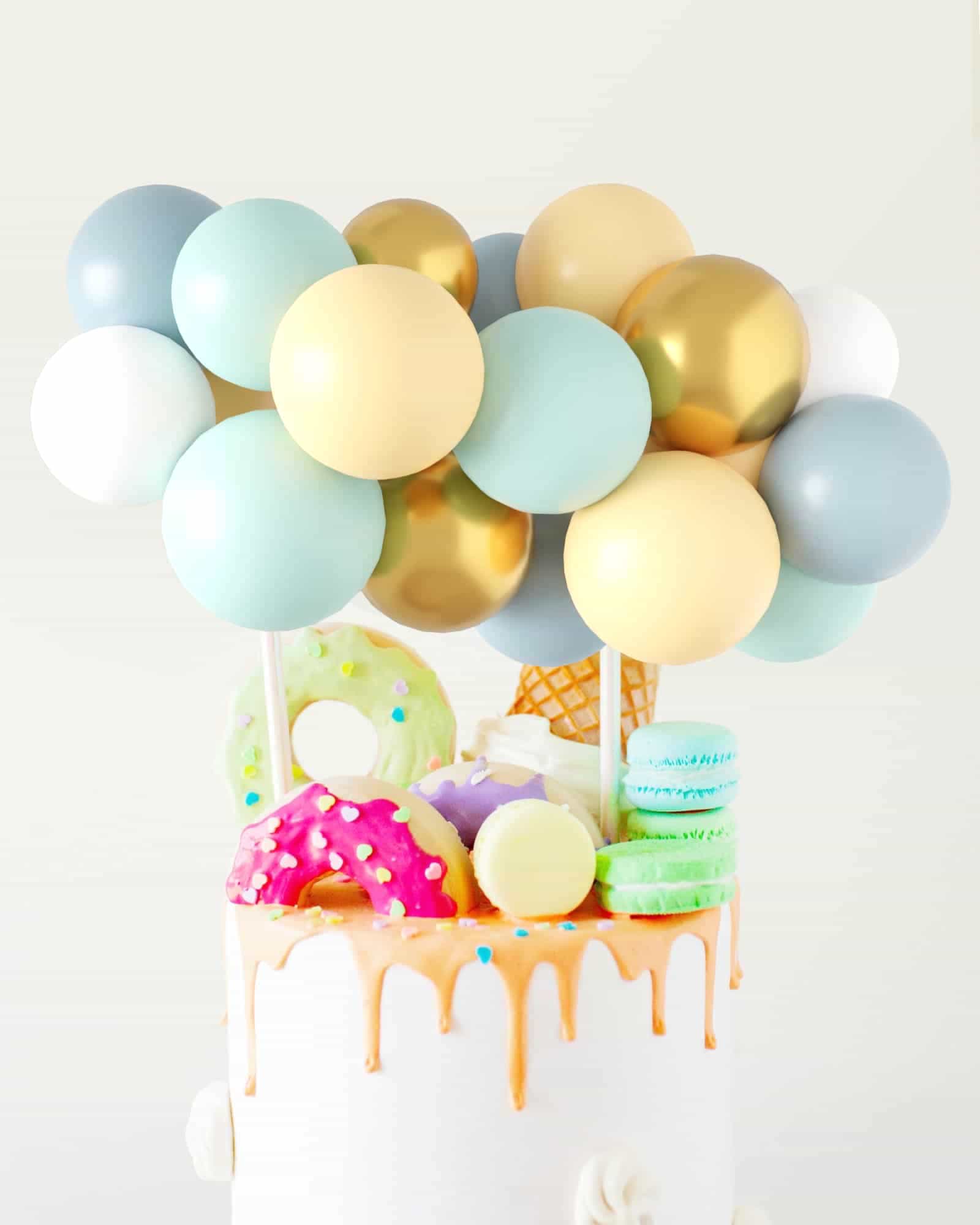 Colorful balloons in shades of blue, green, gold, and cream atop a cake adorned with vibrant sweets and dripping glaze.