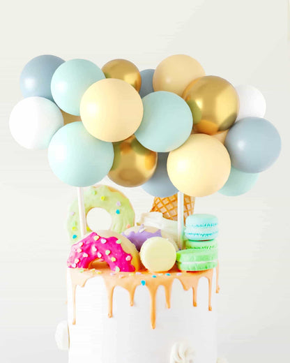 Colorful balloons in shades of blue, green, gold, and cream atop a cake adorned with vibrant sweets and dripping glaze.