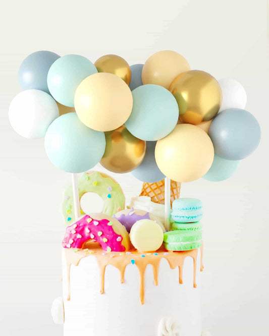 Colorful balloons in shades of blue, green, gold, and cream atop a cake adorned with vibrant sweets and dripping glaze.