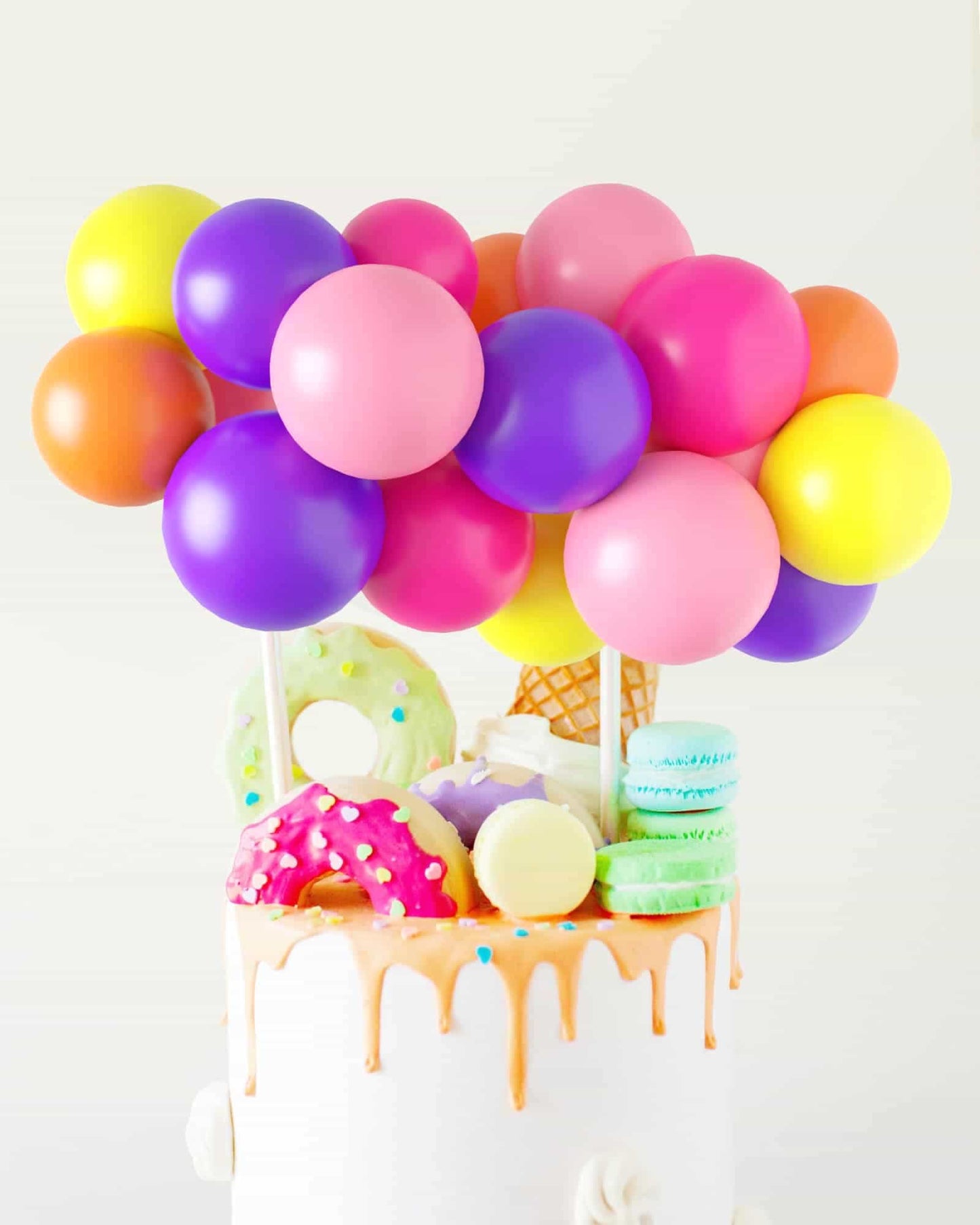 Colorful balloons hover above a cake topped with donuts, macarons, and an ice cream cone, all dripping with frosting.