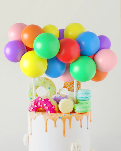 Colorful balloons in various shades hover above a cake topped with vibrant treats like donuts, ice cream, and macarons.