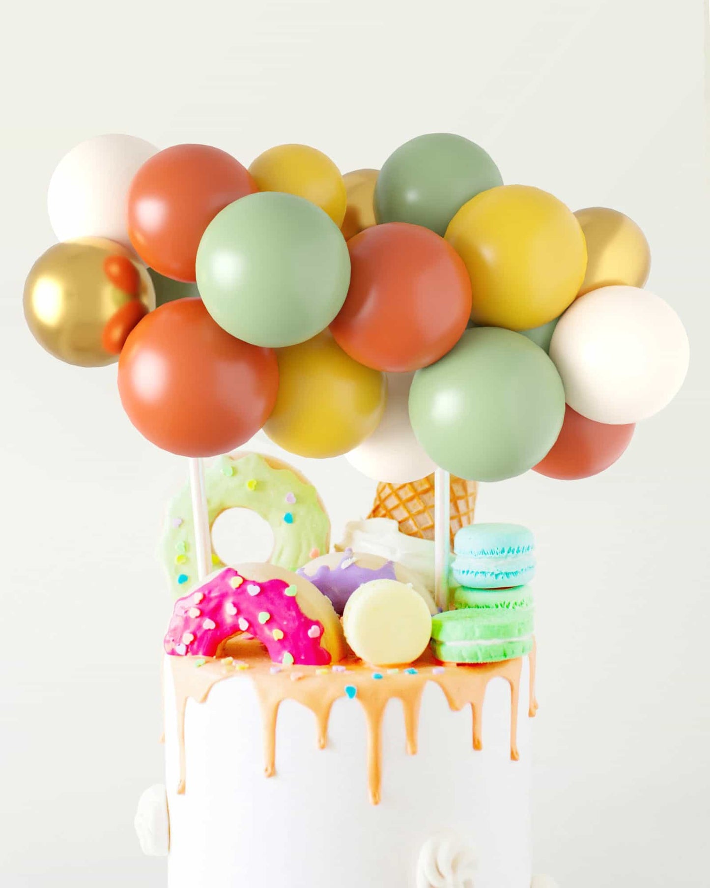 Colorful balloons in shades of green, orange, and gold atop a cake adorned with vibrant treats like donuts and macarons.