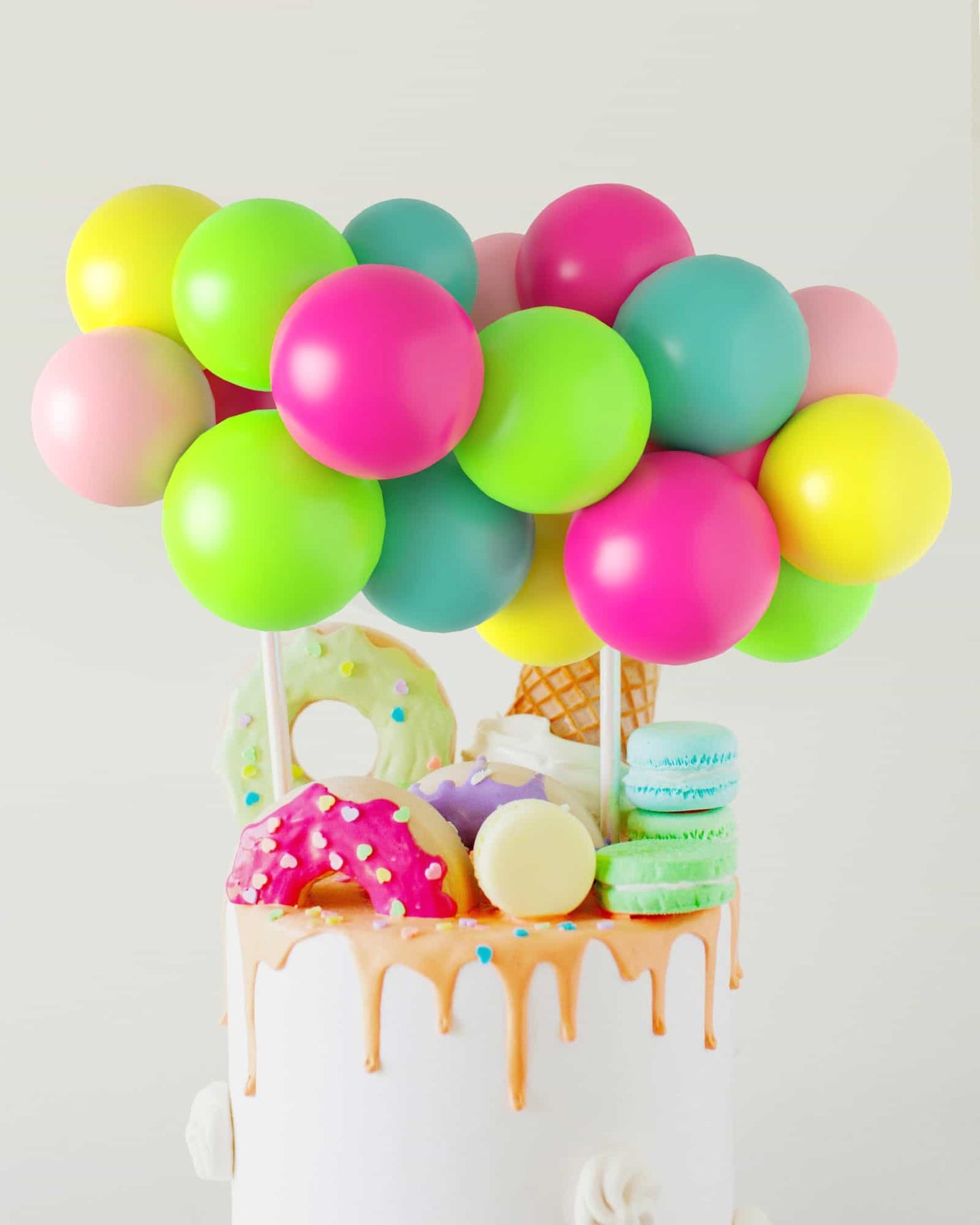 Colorful balloons in vibrant shades hover above a cake adorned with playful sweets and dripping icing.