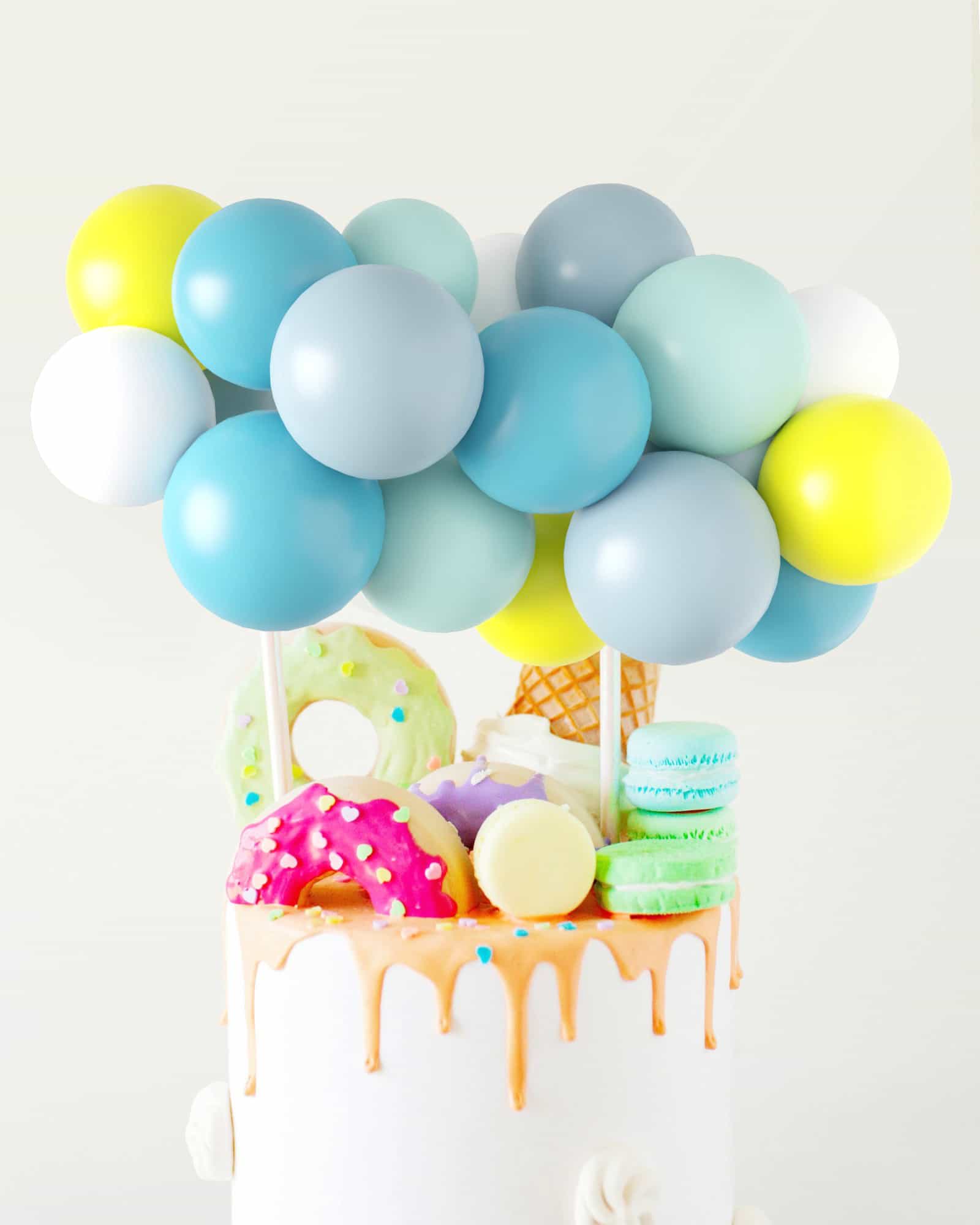 Colorful balloons hover above a festive cake topped with vibrant treats like donuts, macarons, and an ice cream cone.