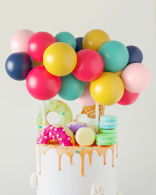 A vibrant cake topped with colorful balloons and playful sweets, featuring donuts, ice cream, and macarons.