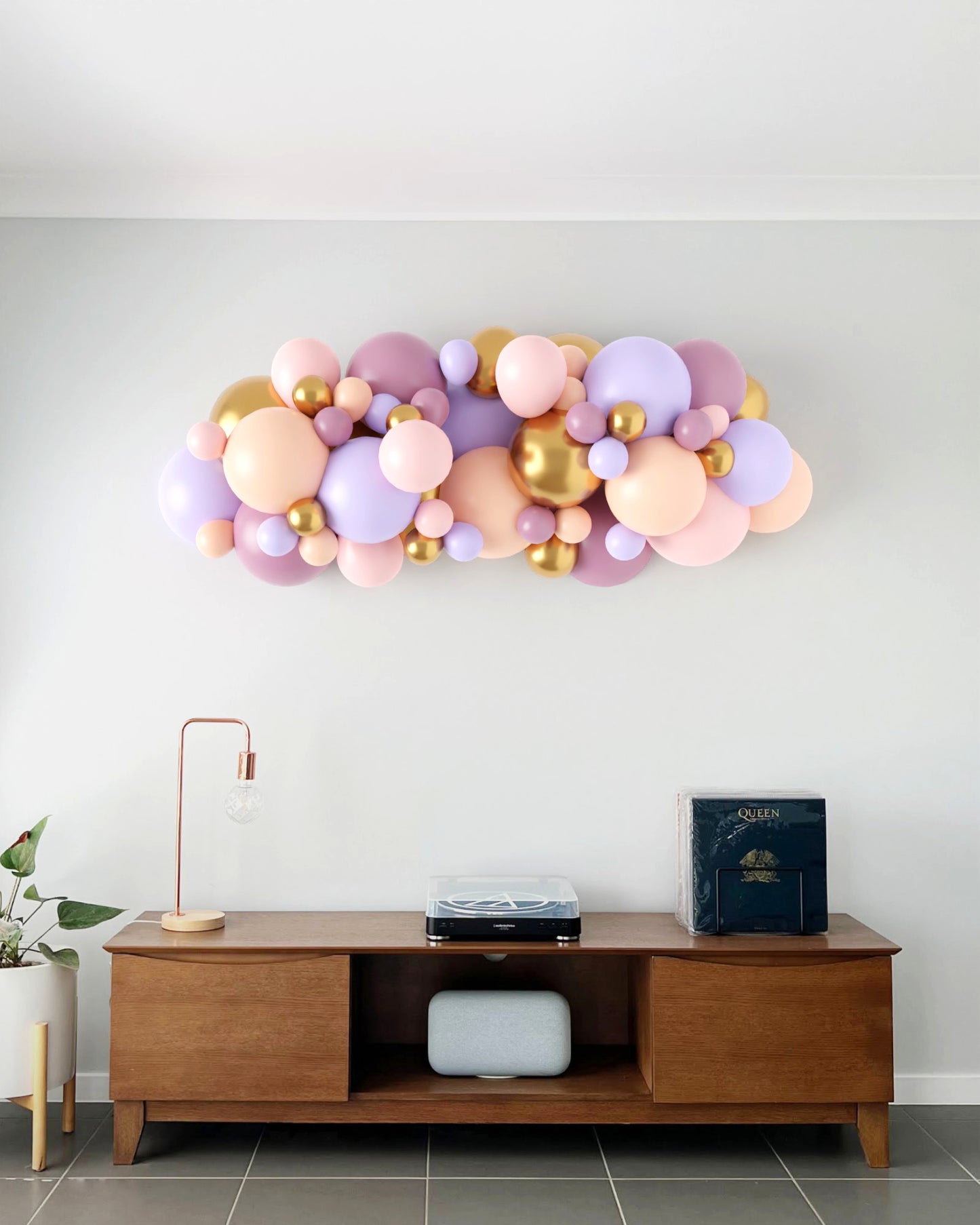 Colorful balloon garland in pastel shades with gold accents, arranged above a wooden console table in a modern setting.