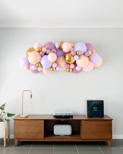 Colorful balloon garland in pastel shades with gold accents, arranged above a wooden console table in a modern setting.
