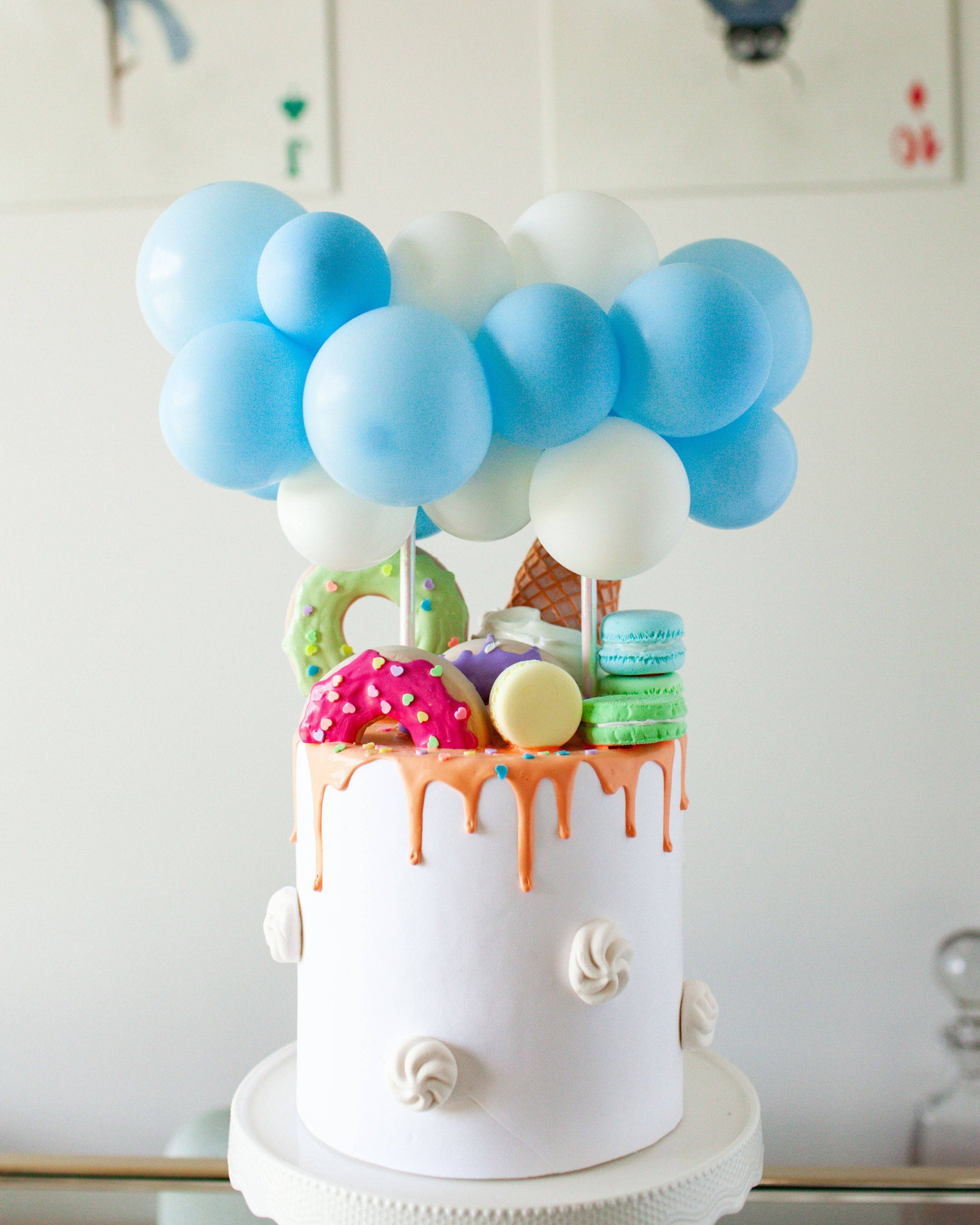 A whimsical cake topped with pastel blue and white balloons, colorful treats, and dripping icing.