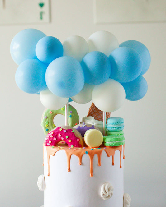 Pastel blue and white balloons atop a whimsical cake adorned with colorful sweets and playful toppings.