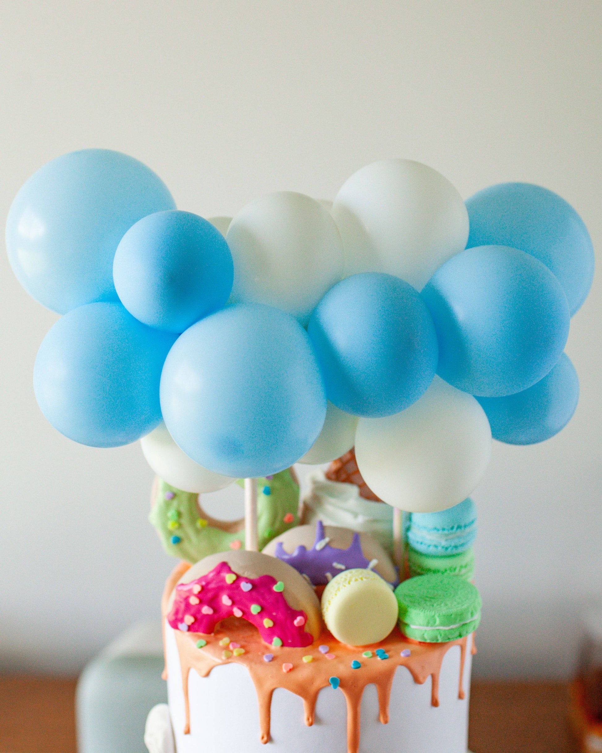 Pastel blue and white balloons atop a colorful, drippy cake adorned with donuts and macarons.