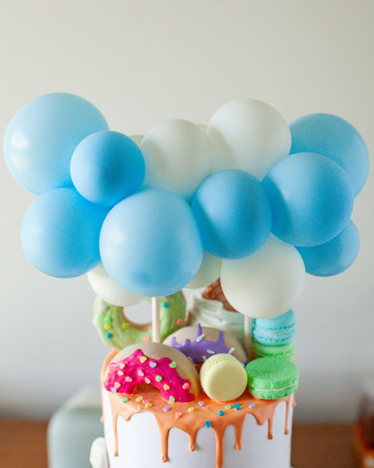 Pastel blue and white balloons atop a colorful, drippy cake adorned with donuts and macarons.