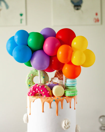 A colorful cake topped with vibrant balloons, sprinkles, donuts, and ice cream cones, creating a festive celebration vibe.