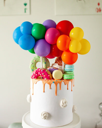 Colorful balloons atop a white cake, adorned with vibrant donuts, macarons, and dripping icing, creating a festive display.