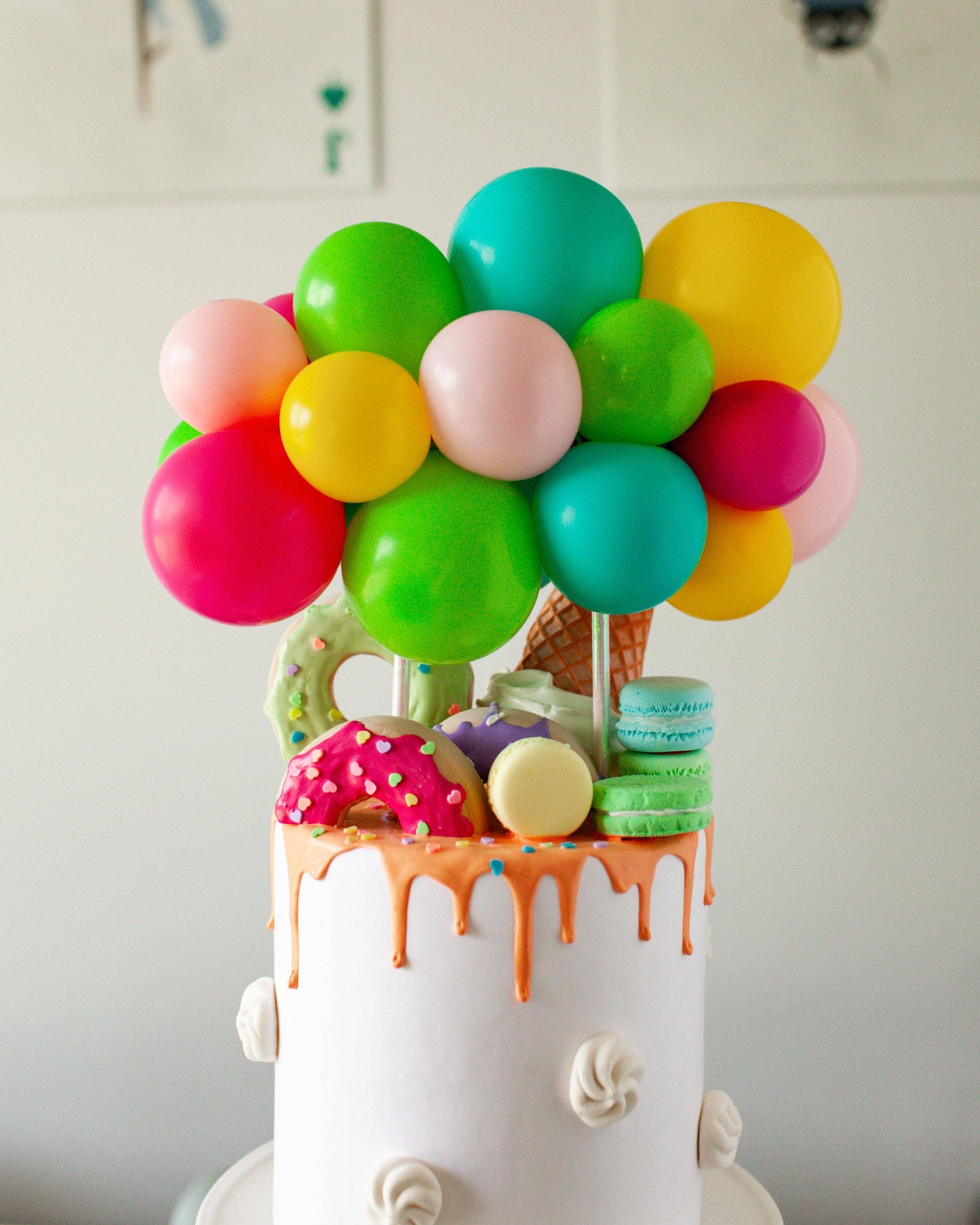 Colorful balloon arrangement atop a decorated cake with dripping icing, donuts, and macarons. Fun and festive display.