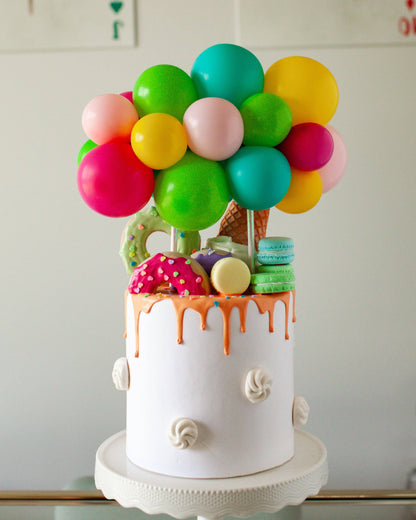 A vibrant cake topped with colorful balloons, donuts, ice cream cones, and macarons, featuring dripping icing.