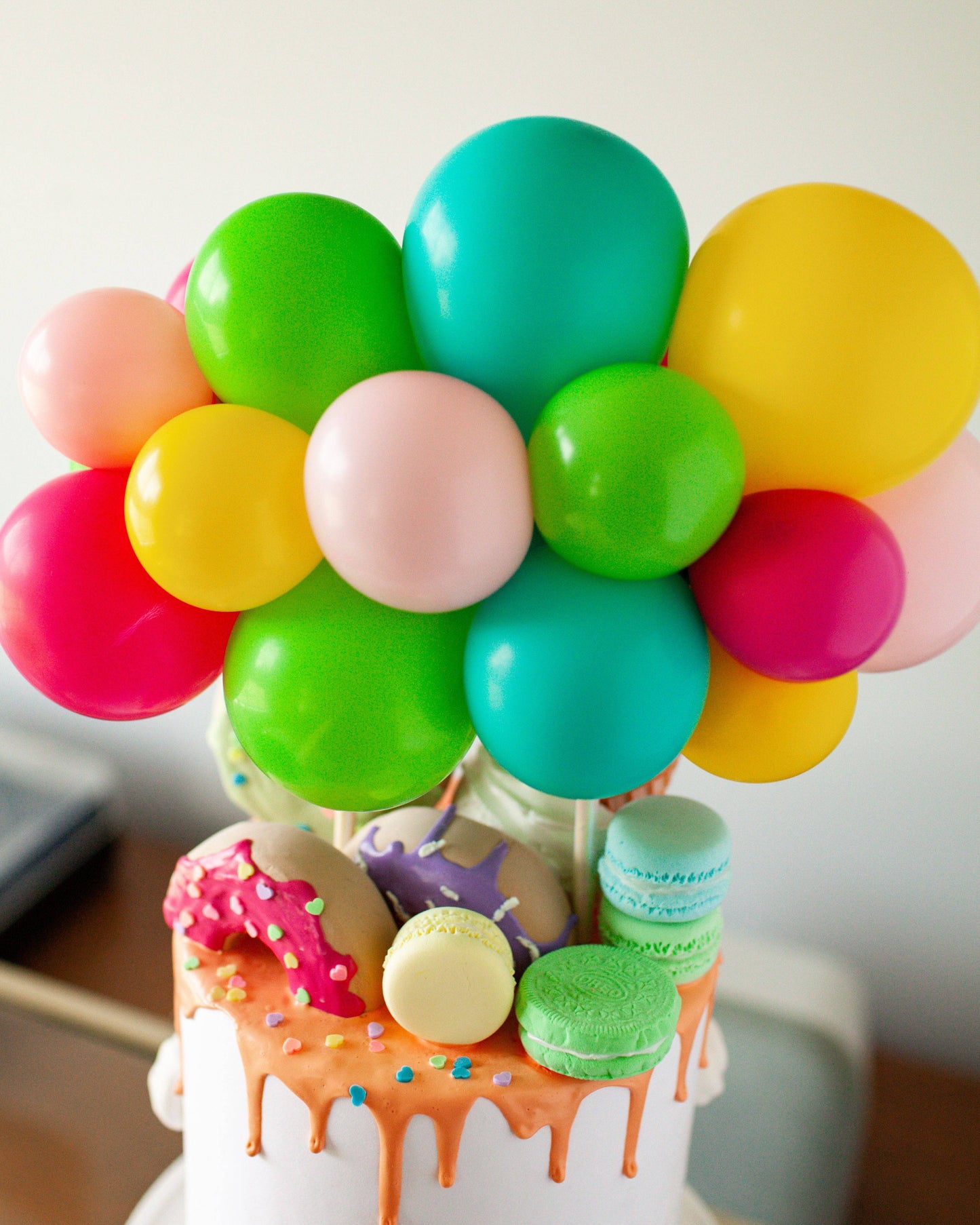 Colorful balloons in vibrant hues topped with decorative macarons and donuts on a drippy frosted cake.