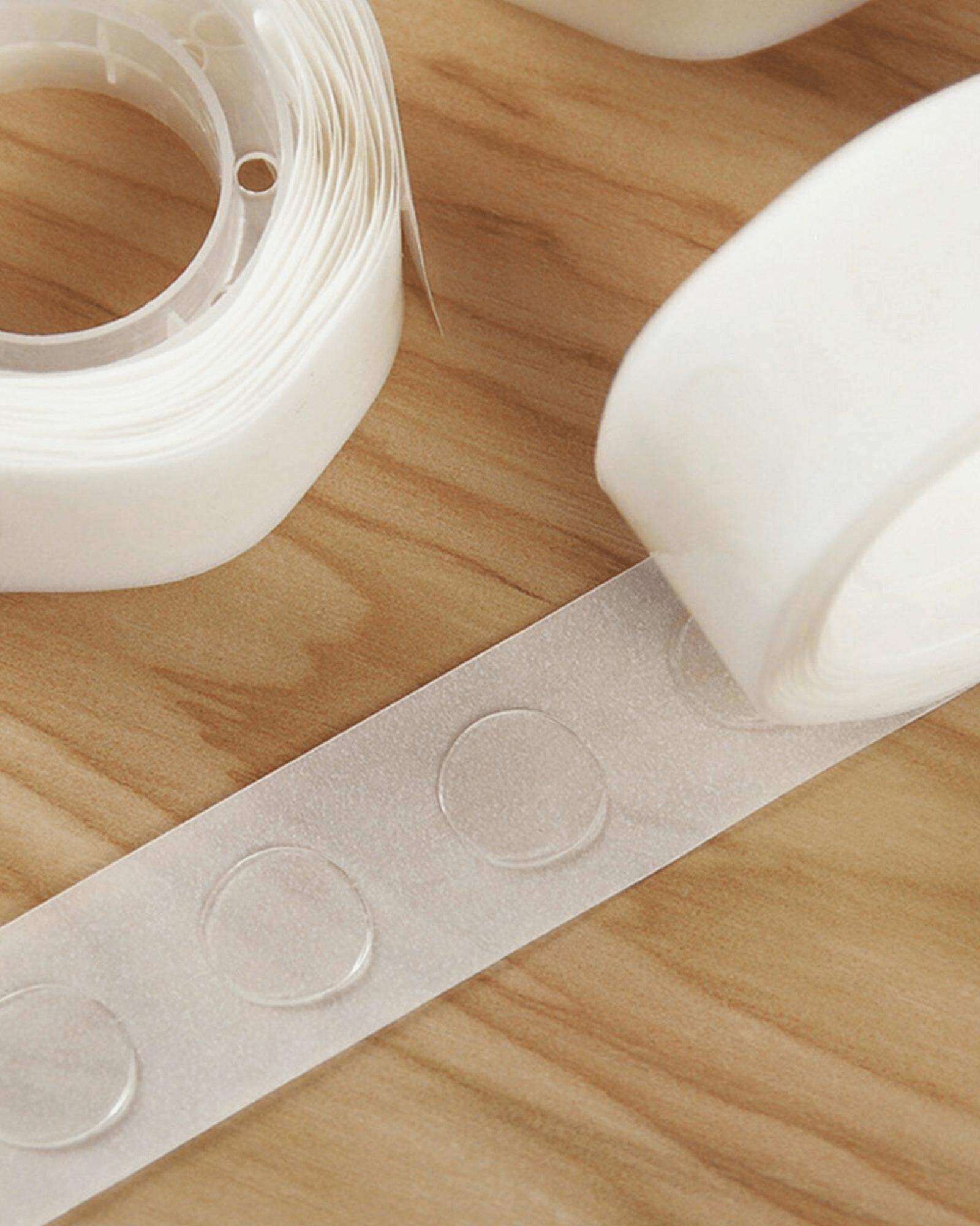 Clear adhesive dots on a wooden surface, with rolls of transparent tape and a smooth strip of adhesive visible.