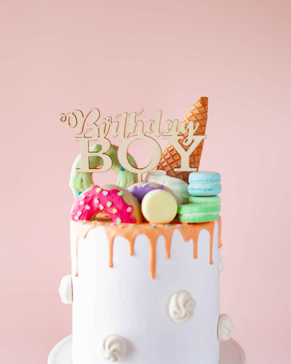 A colorful cake topped with a wooden "Birthday Boy" sign, donuts, macarons, and a cone, against a soft pink backdrop.