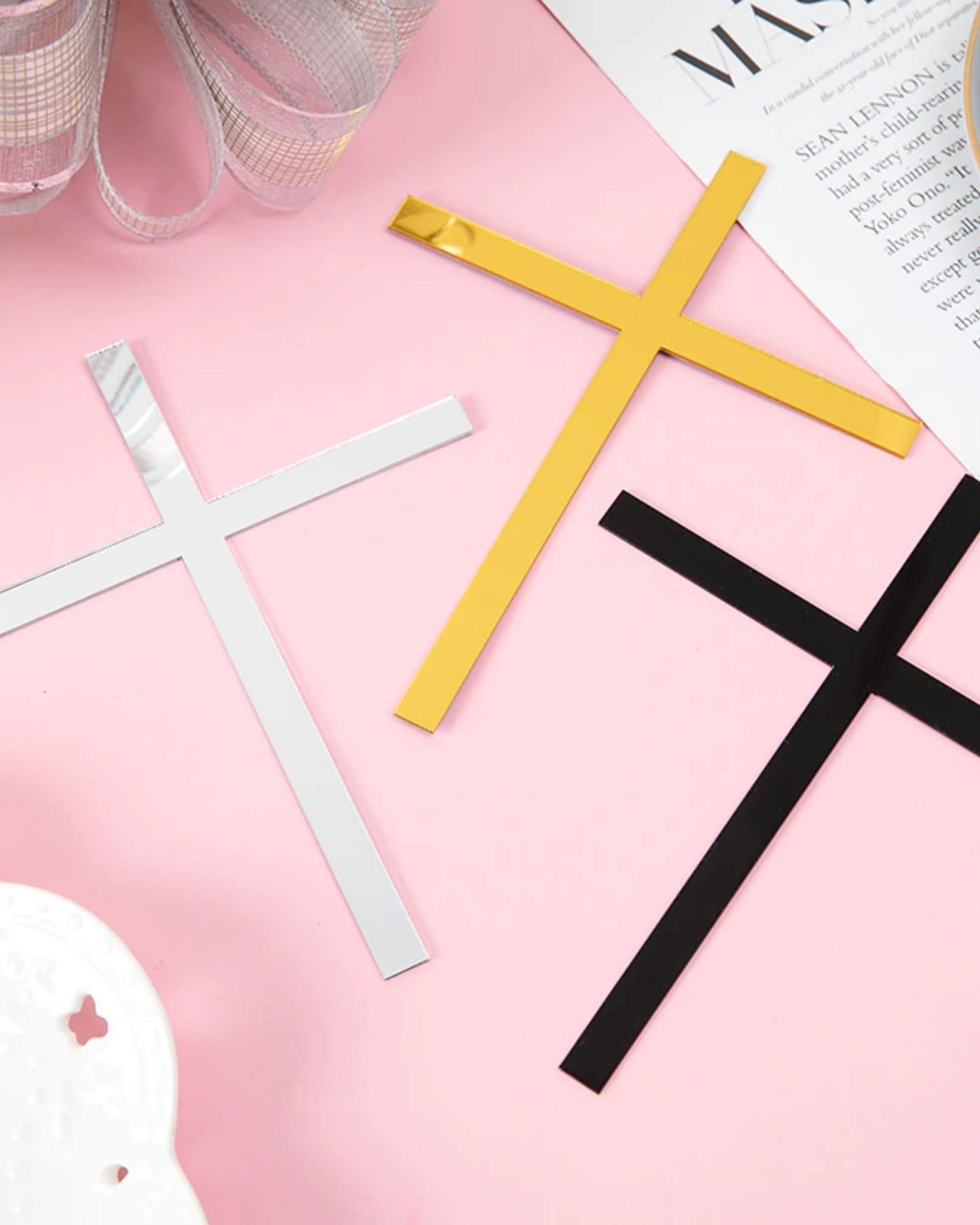 Three cross-shaped decorations in black, white, and gold laid on a soft pink surface alongside a silver ribbon and magazine.