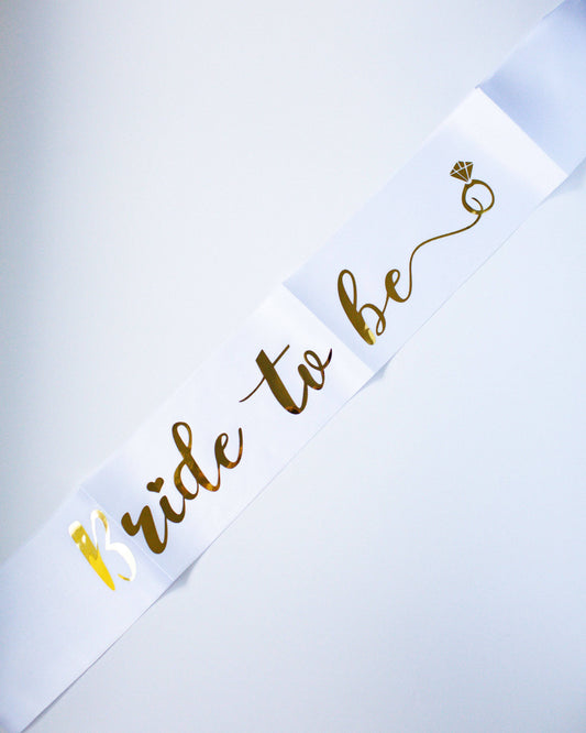 A white sash with elegant gold lettering reading "Bride to Be" and a small diamond icon, perfect for celebrations.