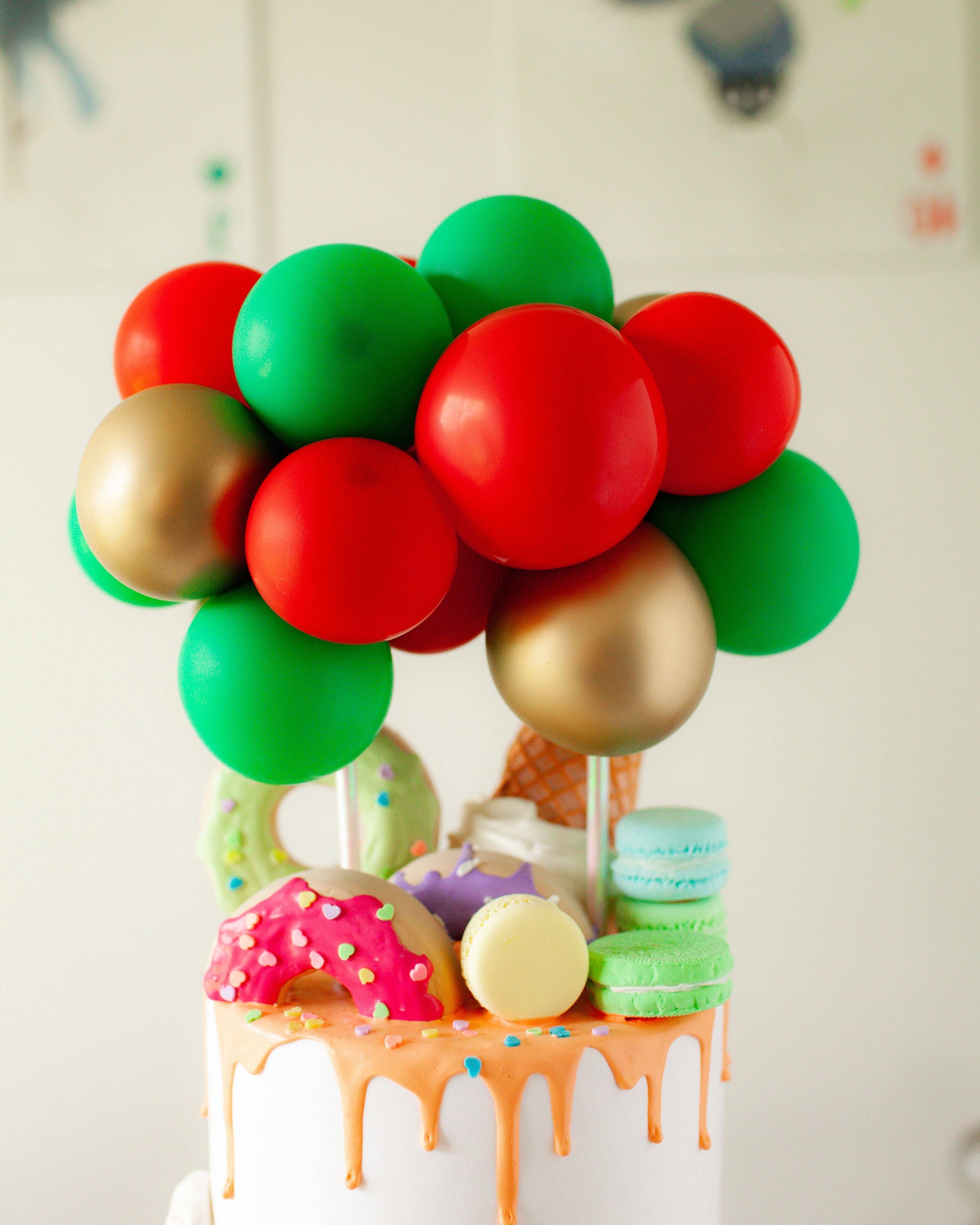 Colorful balloons in red, green, and gold atop a cake decorated with vibrant donuts and macarons, drizzled with orange.