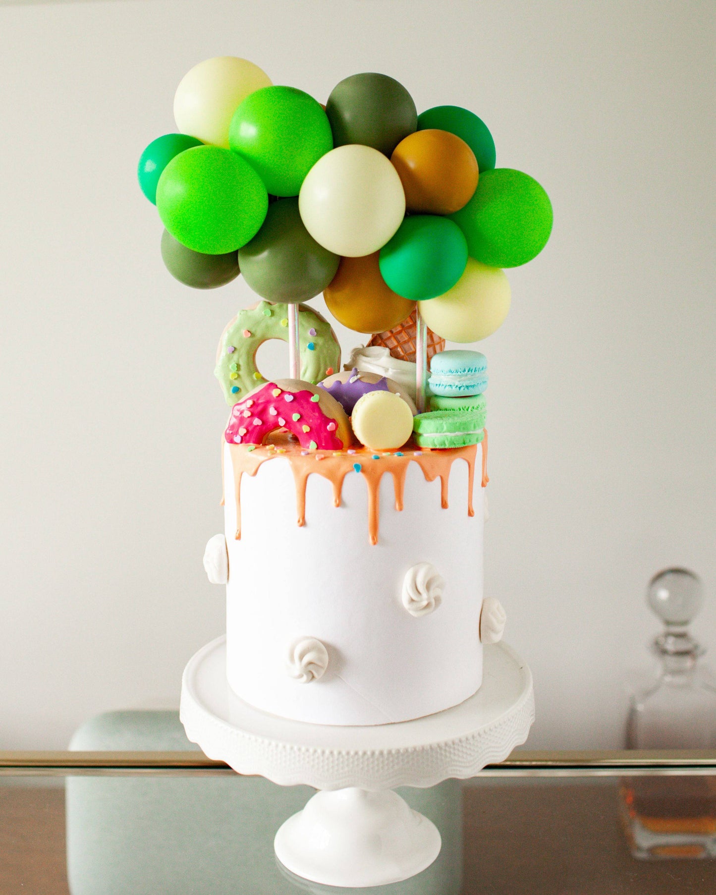 Colorful balloons atop a layered confection, adorned with sweets and sprinkles, on a decorative stand.