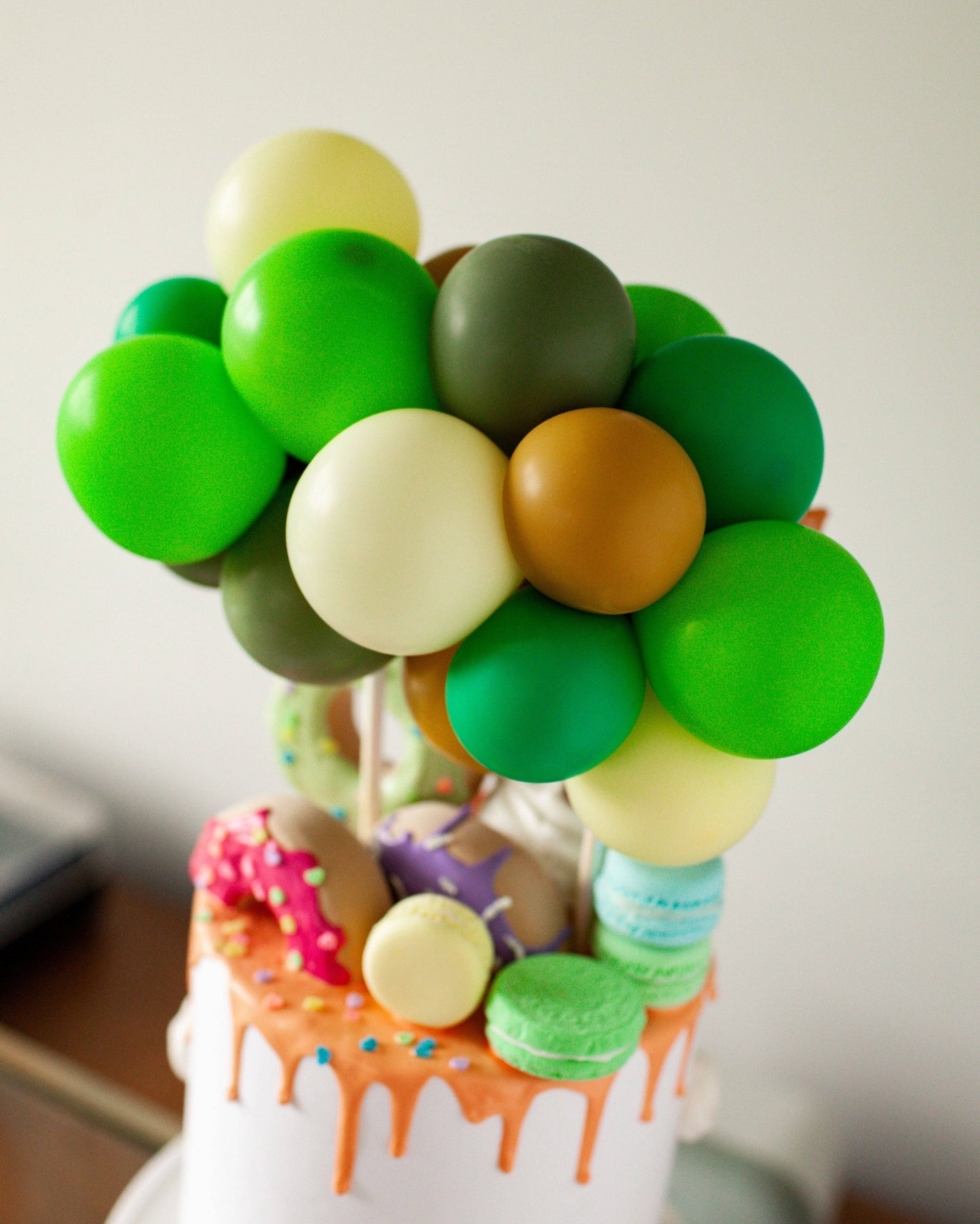 Colorful balloons in various shades of green, brown, and cream atop a cake adorned with sprinkles and macarons.