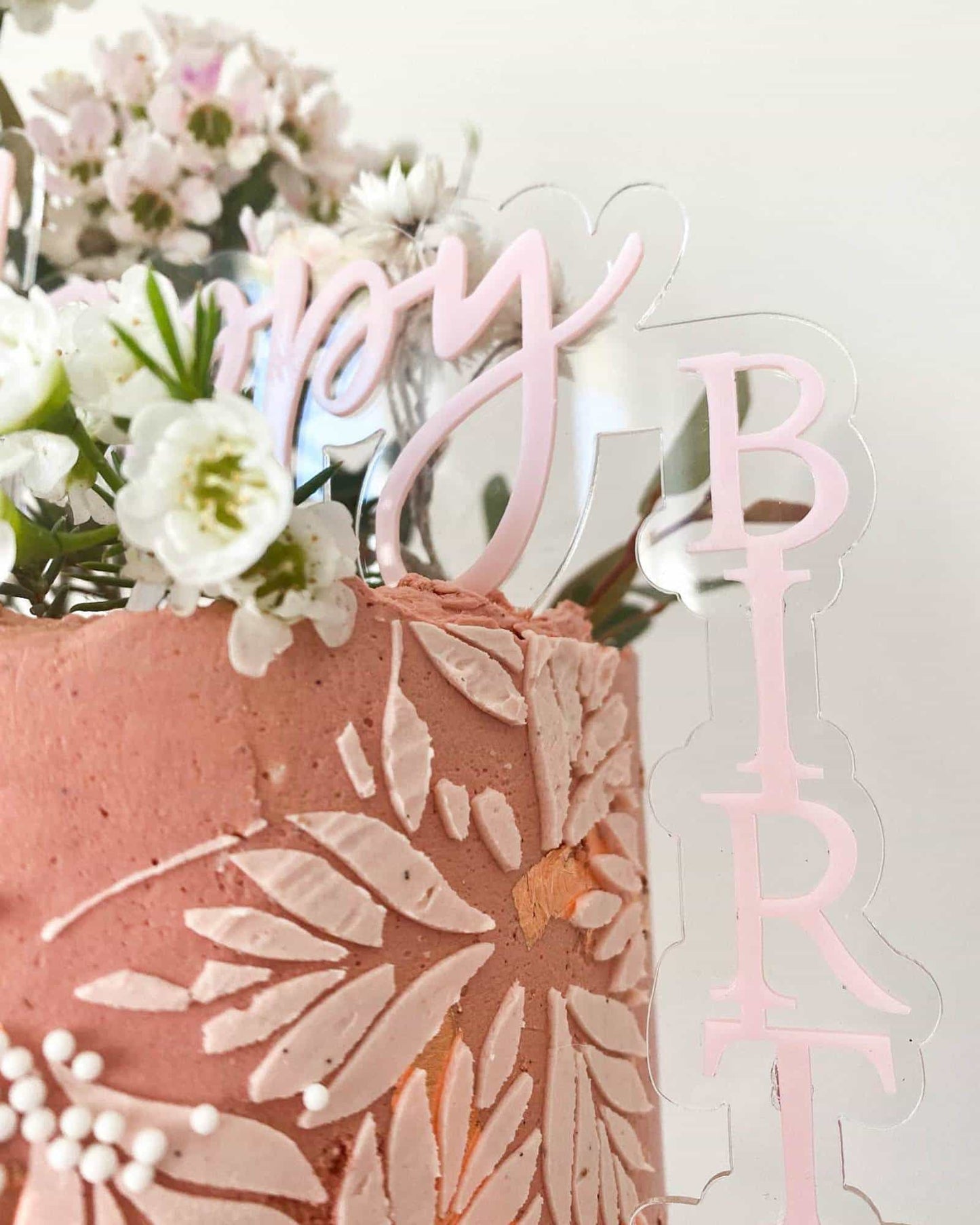 Close-up of a pink cake topped with delicate flowers and a transparent "Happy Birthday" topper.