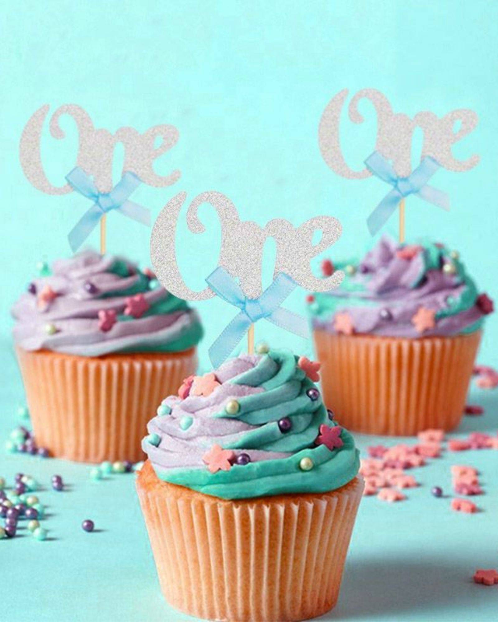 Colorful cupcakes topped with swirls of frosting, adorned with pastel sprinkles and festive "One" picks.