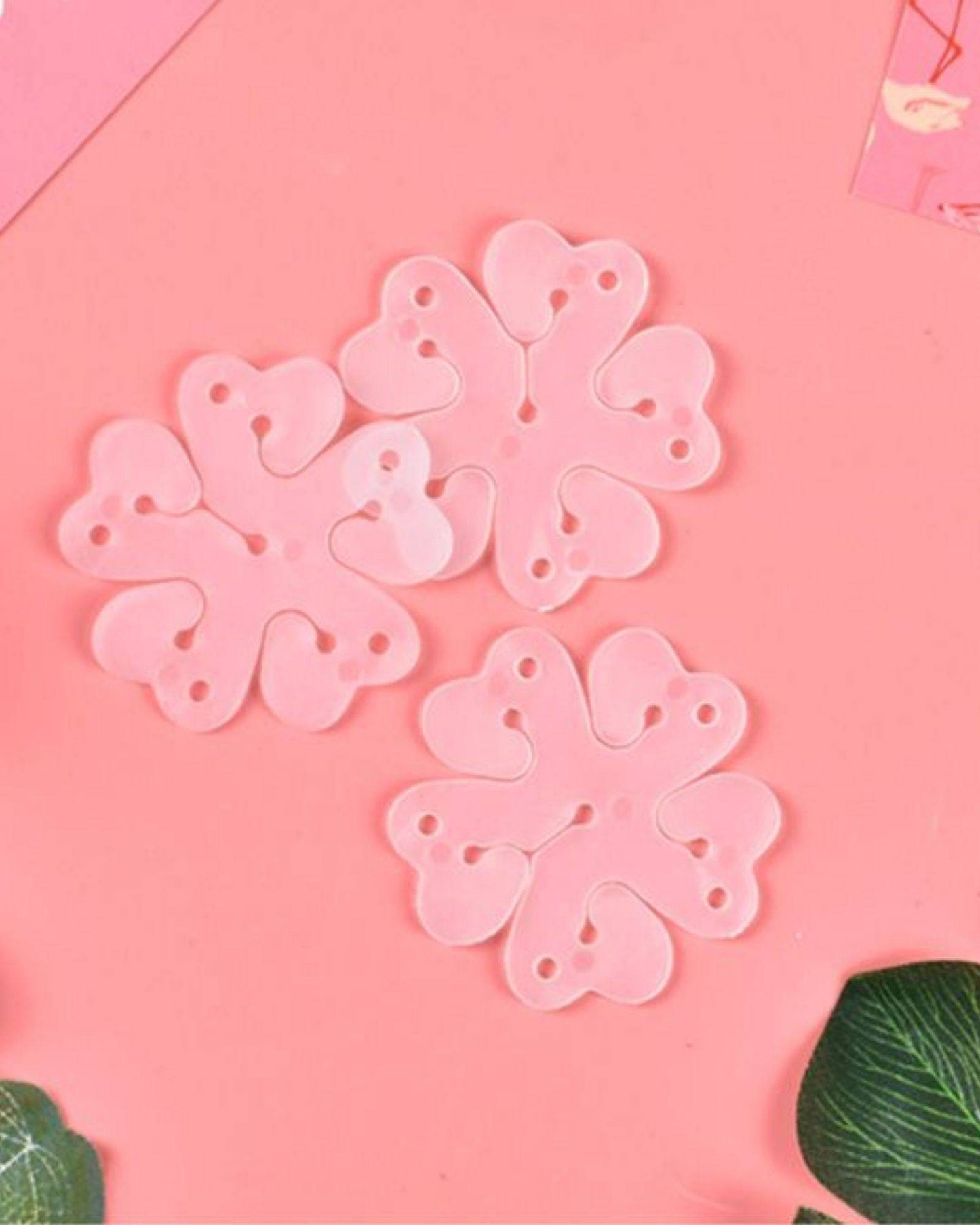 Three pink flower-shaped clips with heart-like petals resting on a soft pink background, surrounded by green leaves.