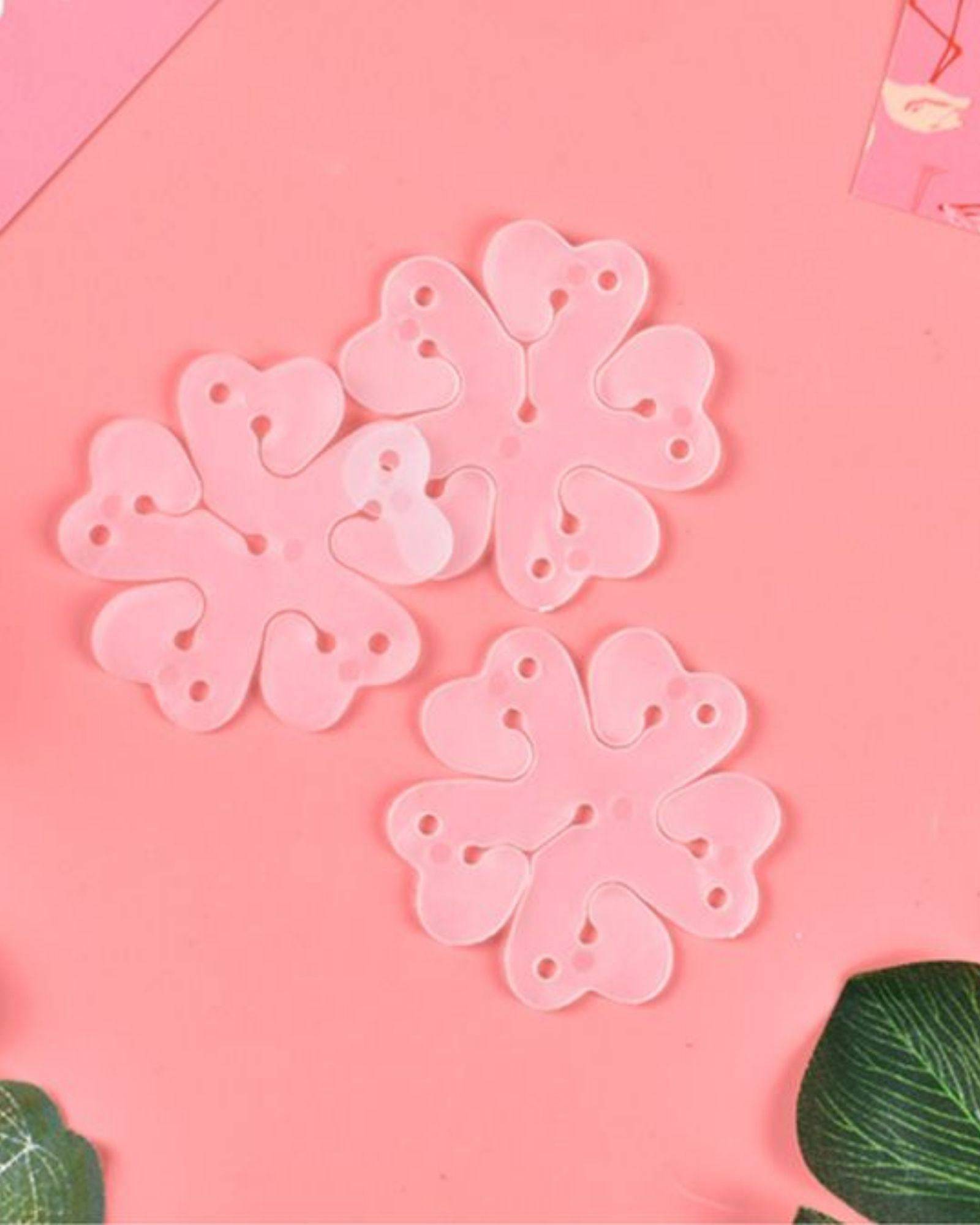 Three pink flower-shaped clips with heart-like petals resting on a soft pink background, surrounded by green leaves.