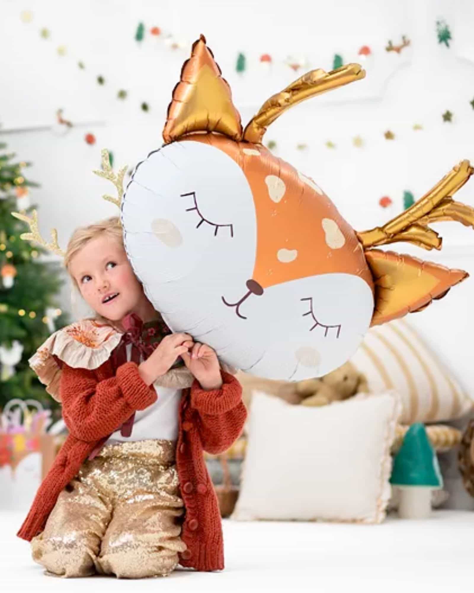 A child in a cozy outfit joyfully holds a large, whimsical deer balloon with golden antlers and a sweet expression.