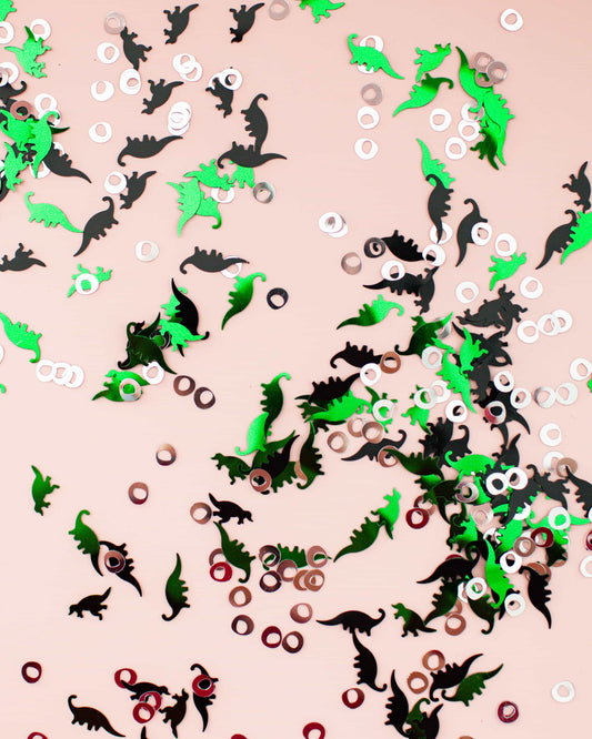 Colorful confetti featuring green and black dinosaur shapes mixed with circular metallic pieces on a light background.