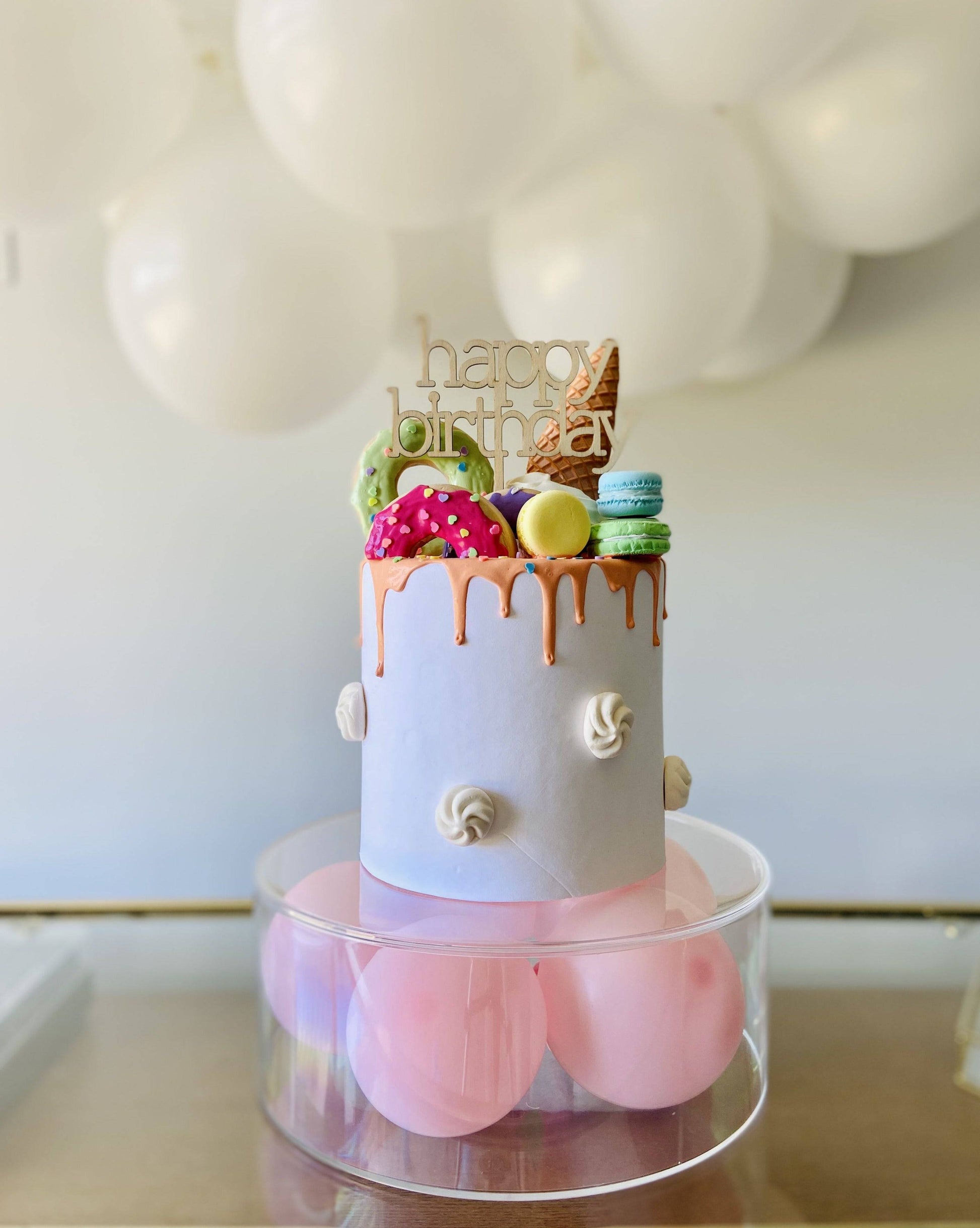 Colorful birthday cake topped with decorative treats, set on a clear base with pink balloons and a festive backdrop.