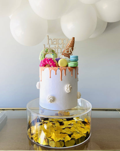 A colorful cake topped with sweets and a "happy birthday" sign, resting on a clear base filled with golden coins.