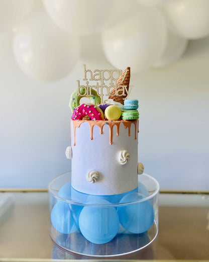 Colorful birthday cake topped with playful decorations, including ice cream and donuts, set on a light blue base.