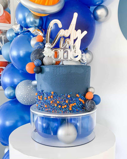 A vibrant blue layered cake adorned with colorful sprinkles and surrounded by festive balloons for a birthday celebration.