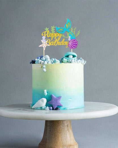 A pastel ombre cake topped with a colorful "Happy Birthday" sign and ocean-themed decorations on a wooden stand.