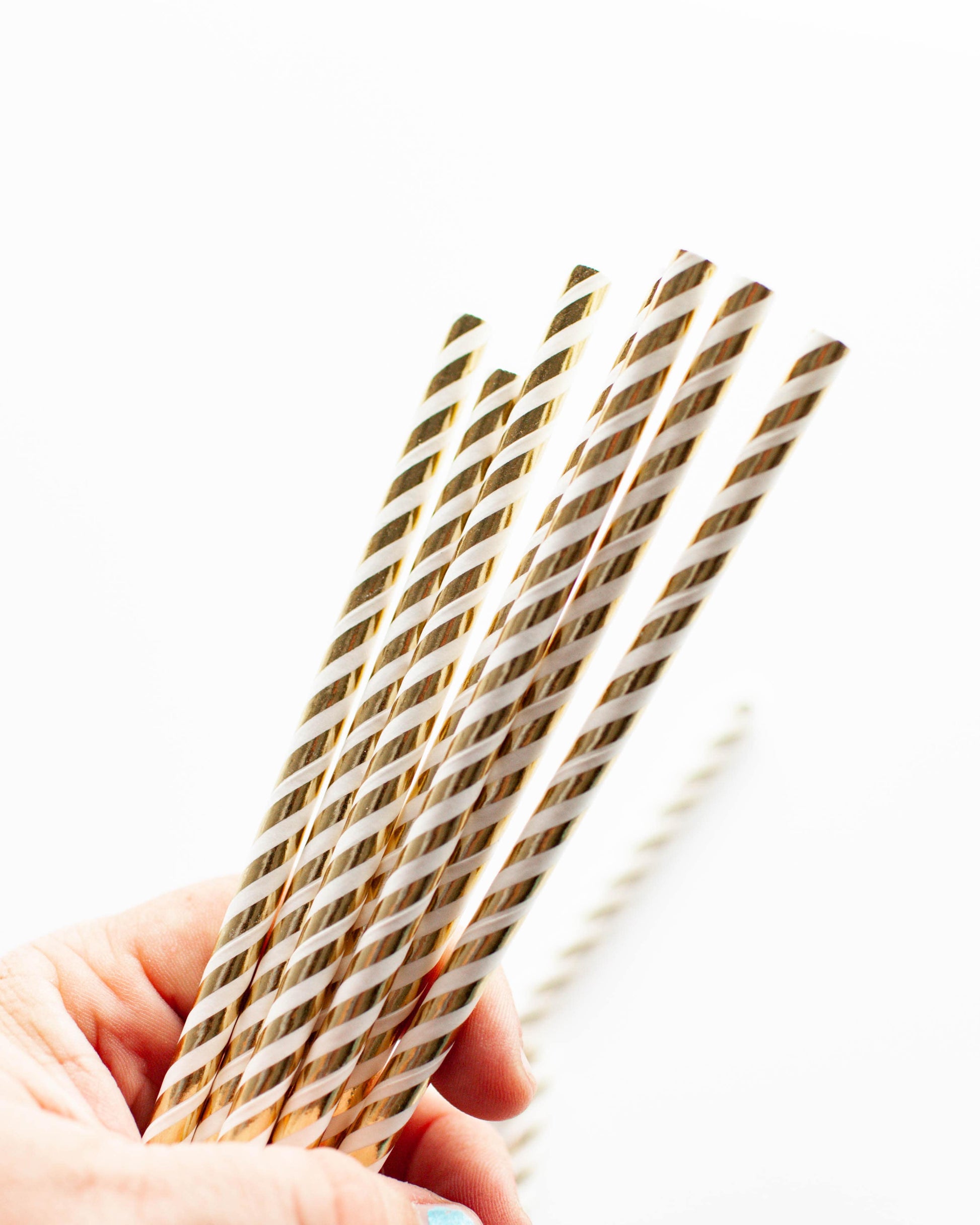 A hand holding several gold foil striped straws against a bright white background.