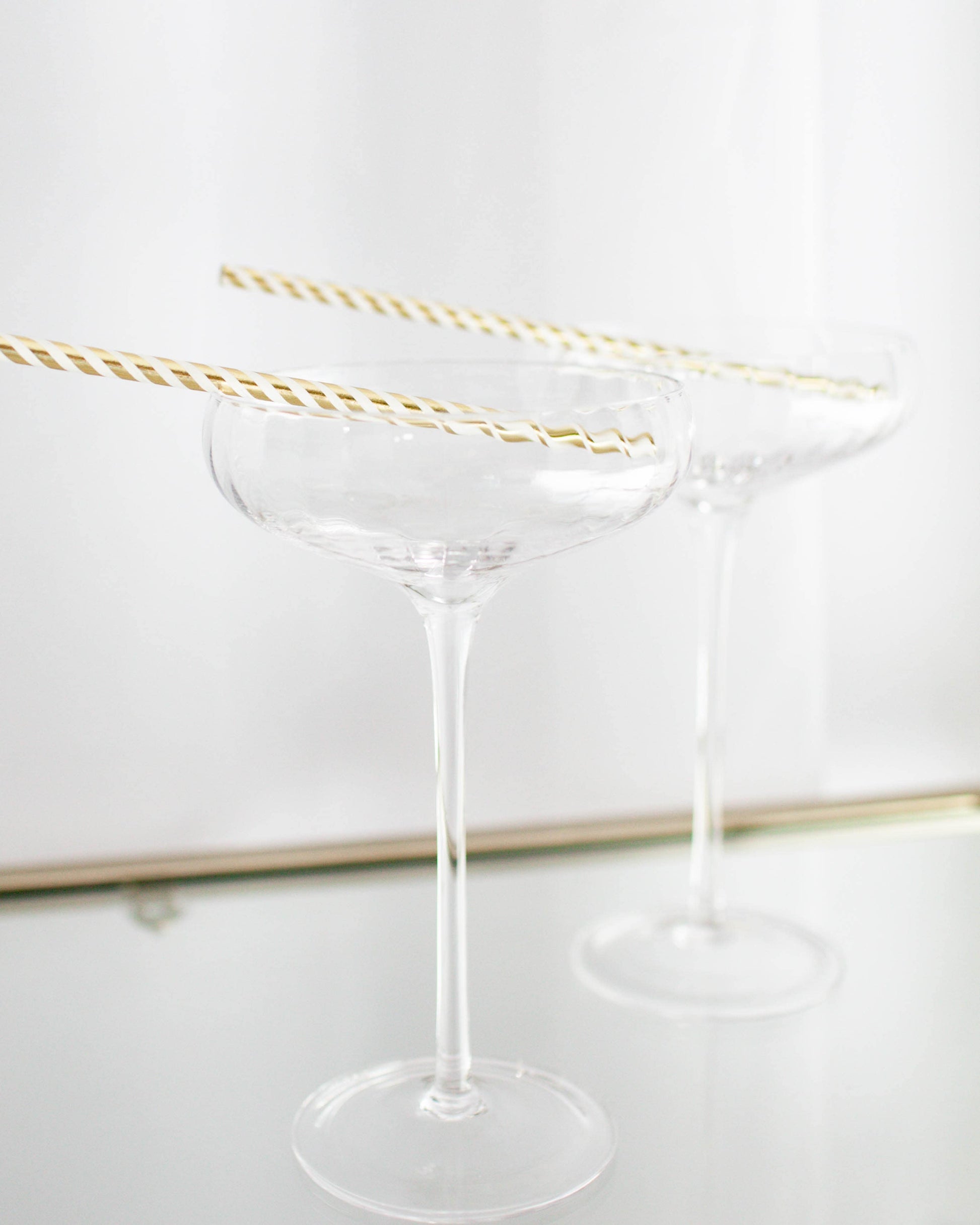 Two clear glass coupe glasses with gold-striped straws resting on the rims against a light background.