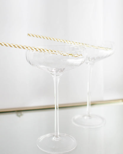 Two clear glass coupe glasses with gold-striped straws resting on the rims against a light background.