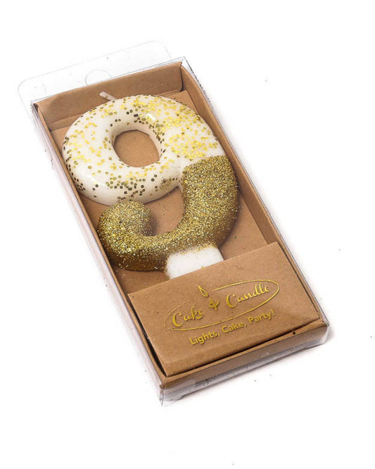 Gold glitter-dipped candle shaped like the number 9, resting in a clear box with a brown base.