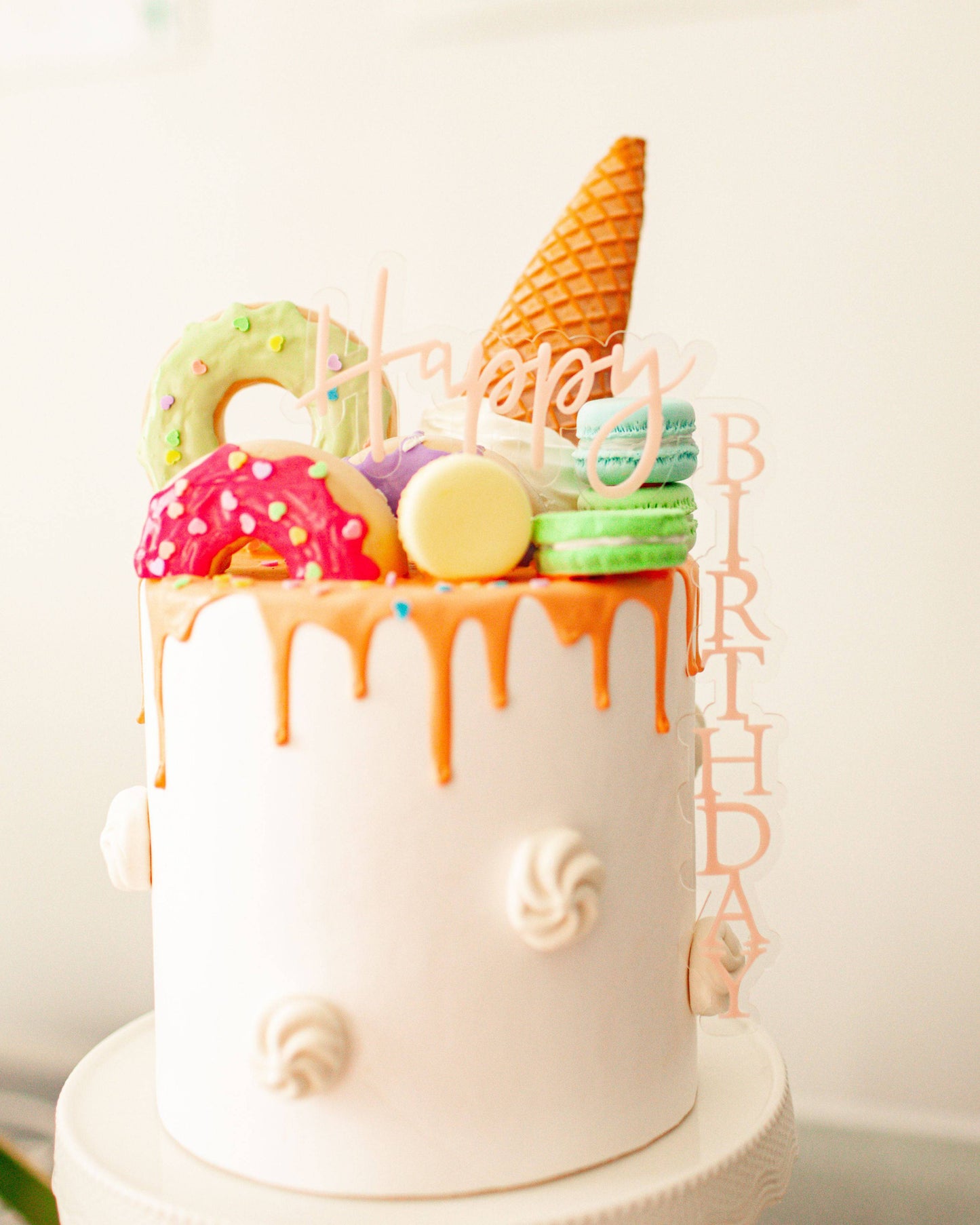 Colorful cake topped with donuts, macarons, and a waffle cone, featuring a "Happy Birthday" topper in pastel hues.