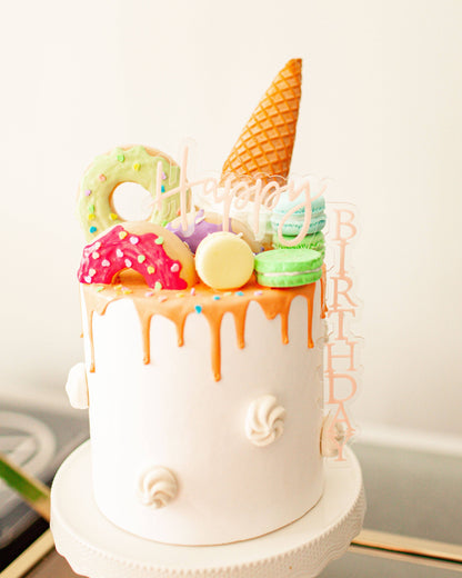 A colorful birthday cake topped with macarons, donuts, and an ice cream cone, featuring a "Happy Birthday" decoration.