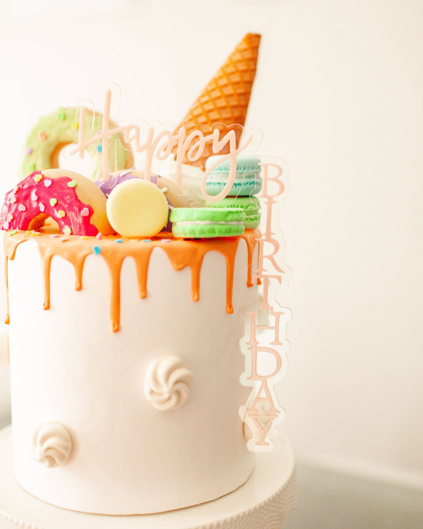 A colorful cake topped with a waffle cone, donuts, macarons, and a festive "Happy Birthday" decoration.