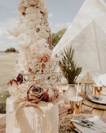 A wooden "Happy Birthday" topper adorned with dried roses and delicate flowers, set against a festive outdoor backdrop.