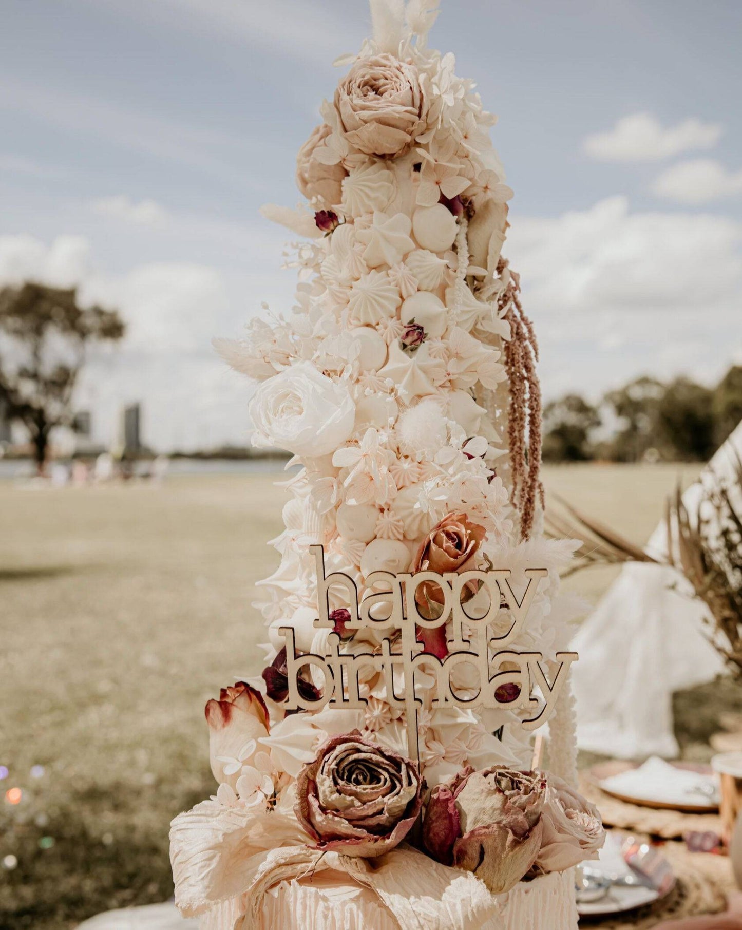 Elegant cake topper adorned with soft pastel flowers and greenery, featuring a wooden "happy birthday" inscription.