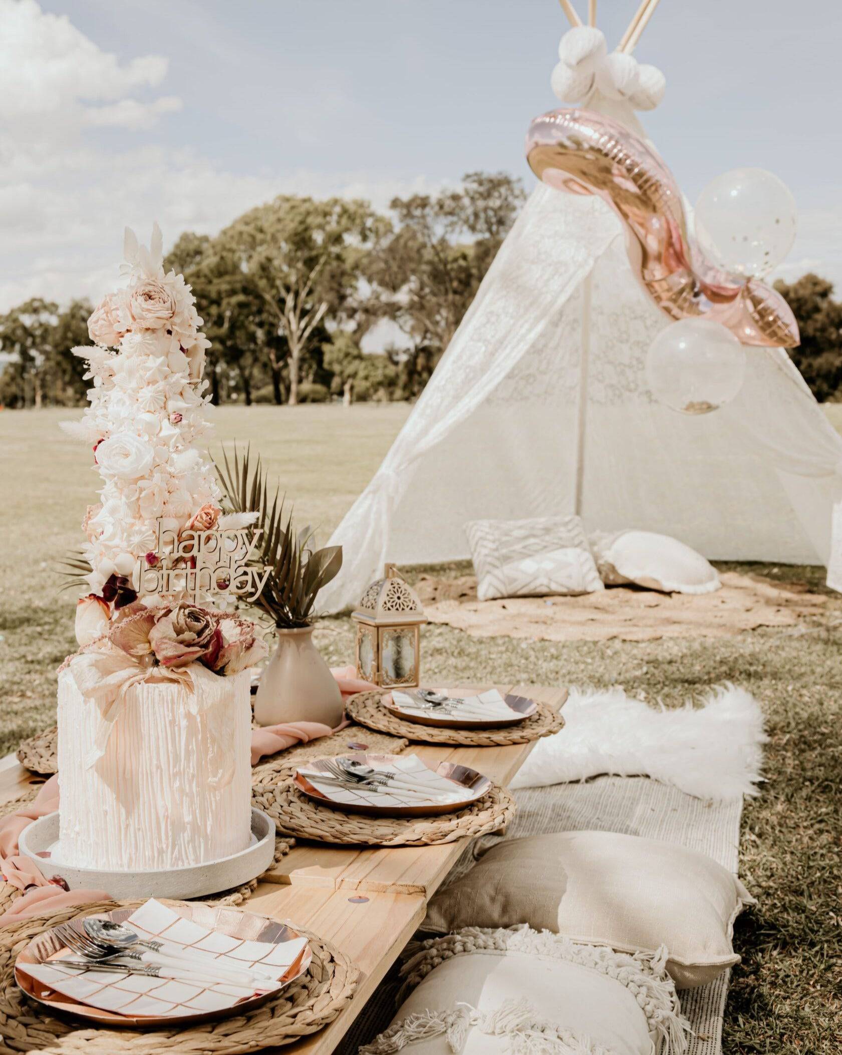 A beautifully arranged picnic scene featuring a birthday cake topped with flowers, elegant plates, and cozy cushions.